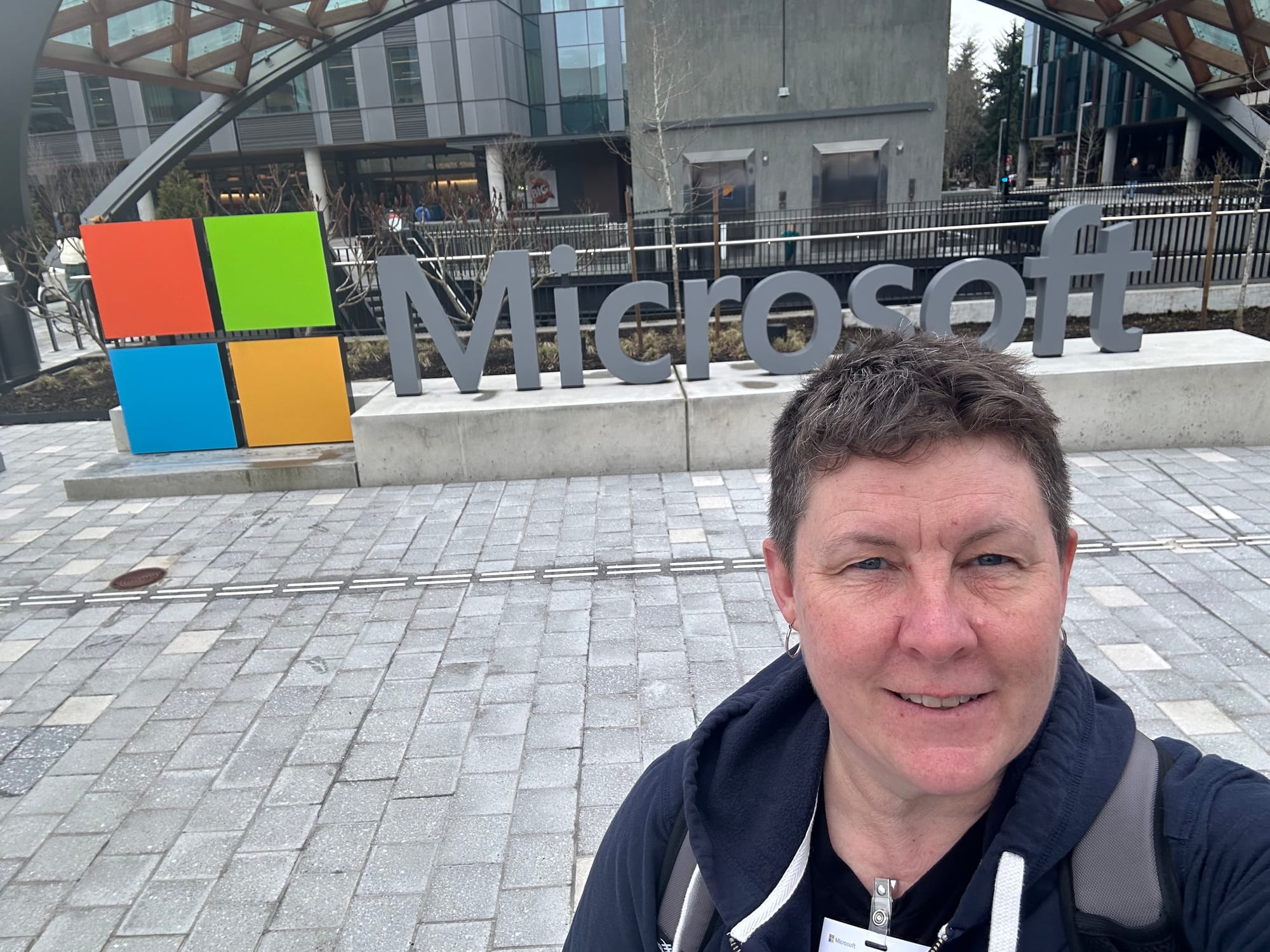 Selfie of me in a blue hooded sweatshirt, short brown hair, standing on a stone sidewalk in front of a Microsoft logo and sign.