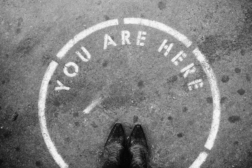 person wearing black leather shoes in front of a sign on the ground that says "You Are Here".