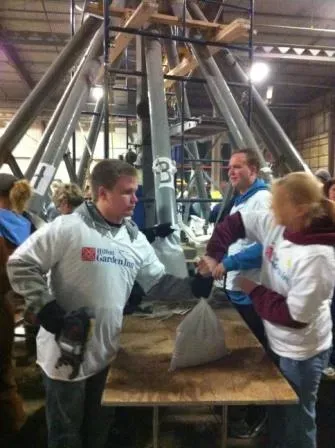 A pic of the "spider" filling machine. Three visible people all in white t-shirts, in the background a large machine with several "legs" where sand is coming out of to fill the bags.
