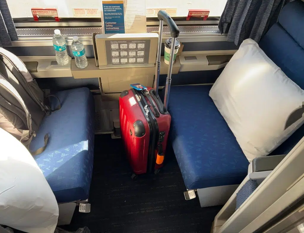 Amtrak roomette interior with two blue bench seats, white pillows, red rolling suitcase, water bottles, and reading lamp.