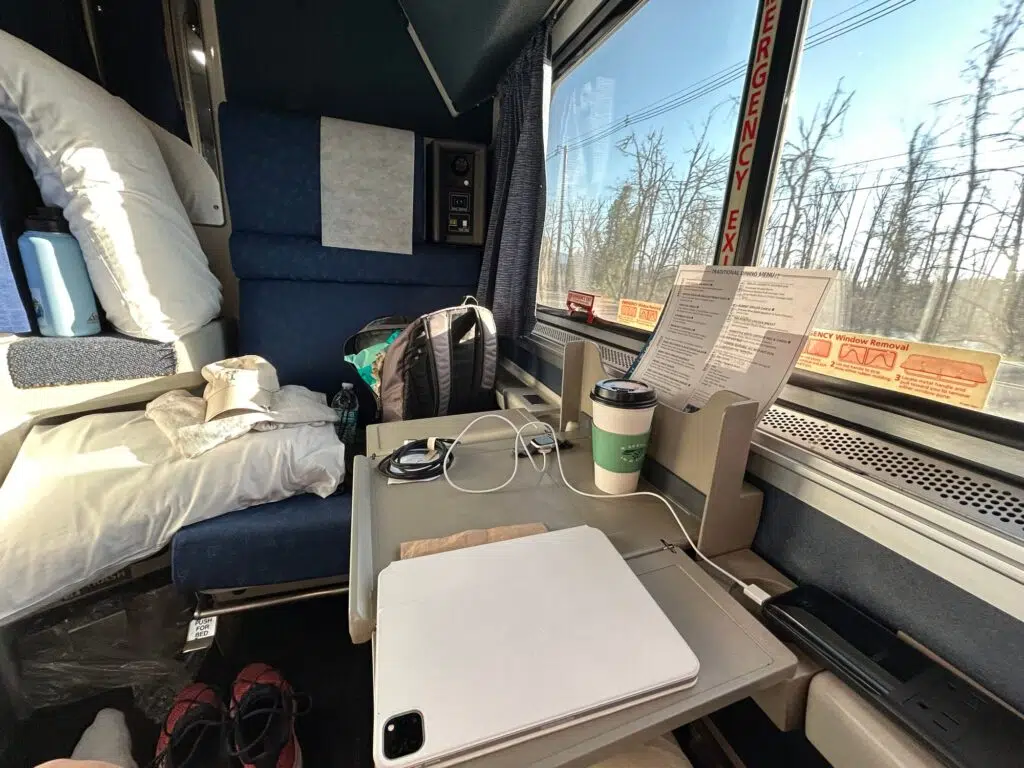 Amtrak roomette interior with blue seats, fold-down table, laptop, coffee cup, charging cables, and window view of trees.