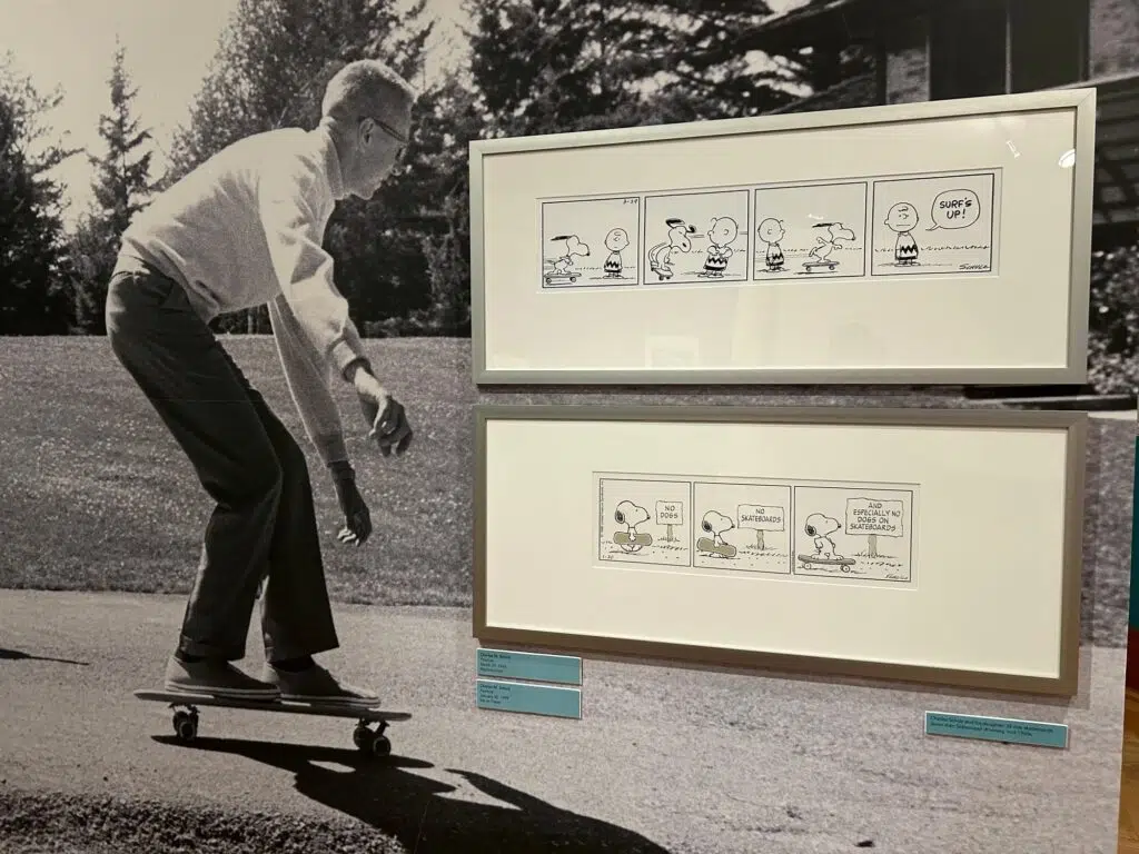 Black and white photo of Charles Schulz skateboarding alongside two framed Peanuts comic strips with Snoopy on a skateboard.