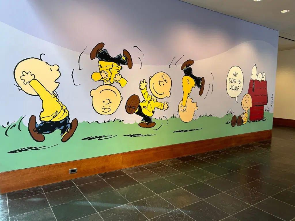 Peanuts mural showing Charlie Brown doing flips across a grassy field, ending at Snoopy's doghouse with speech bubble "My dog is home!".