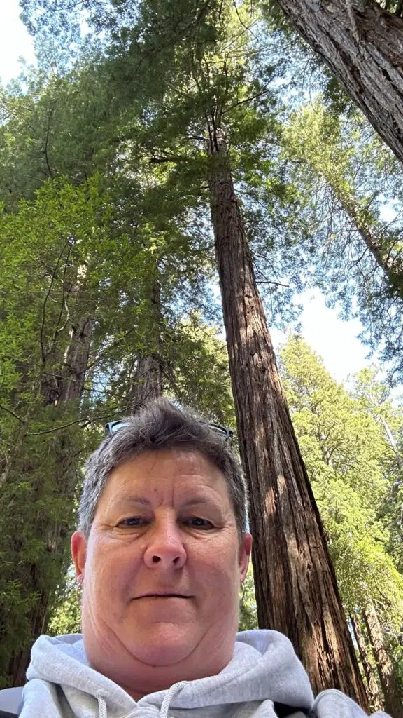 Me taking selfie among towering redwood trees at Muir Woods National Monument, sunlight filtering through green canopy.