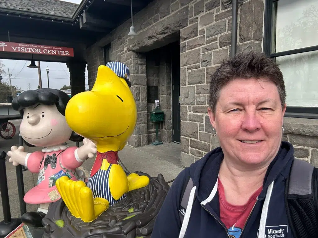 Me in a Microsoft jacket taking selfie with Peanuts character statues of Lucy and Woodstock at Santa Rosa Visitor Center.